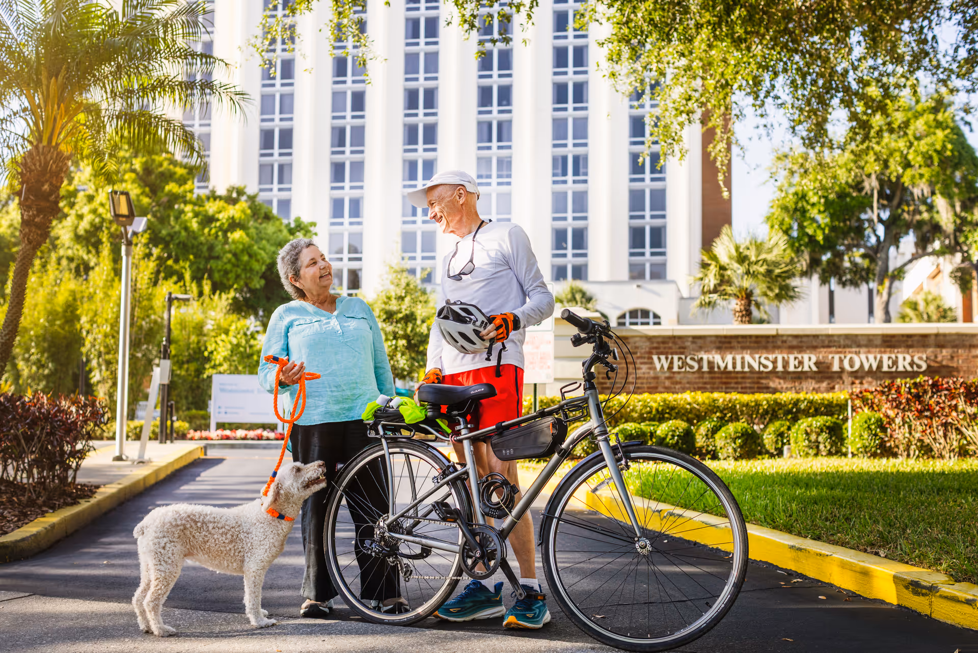 An elderly man and woman standing outside Westminster Towers. The man is holding a bicycle and wearing a white cap, white long-sleeve shirt, red shorts, and gloves. The woman is holding a leash attached to a small white dog. They are smiling and engaging in conversation with greenery and the building in the background.