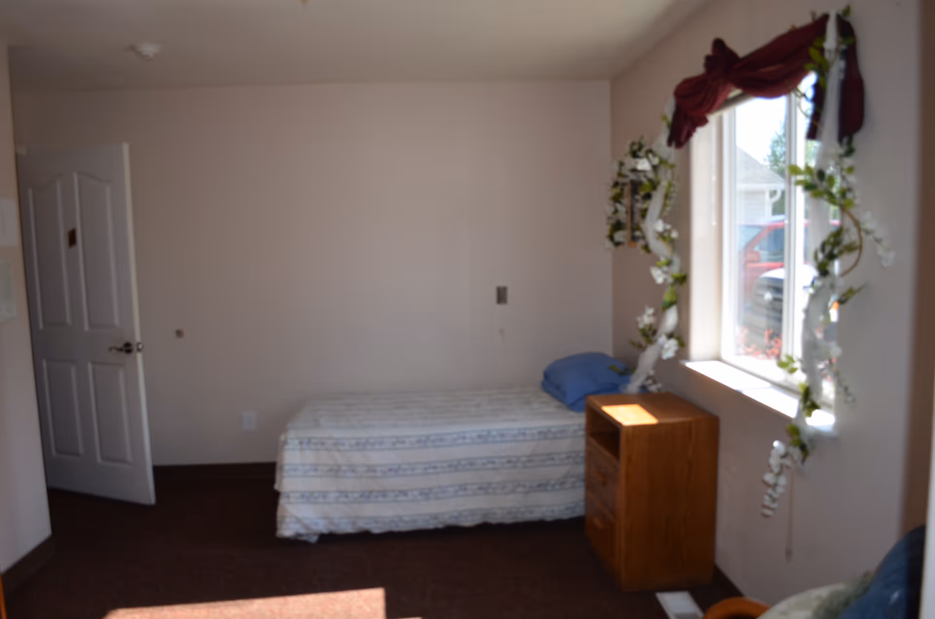 A small bedroom with a single bed covered with a patterned bedspread and a blue pillow. Next to the bed is a wooden nightstand with three drawers. A window with floral garlands and a burgundy valance lets in natural light. The room has beige walls, a brown carpet, and an open white door.
