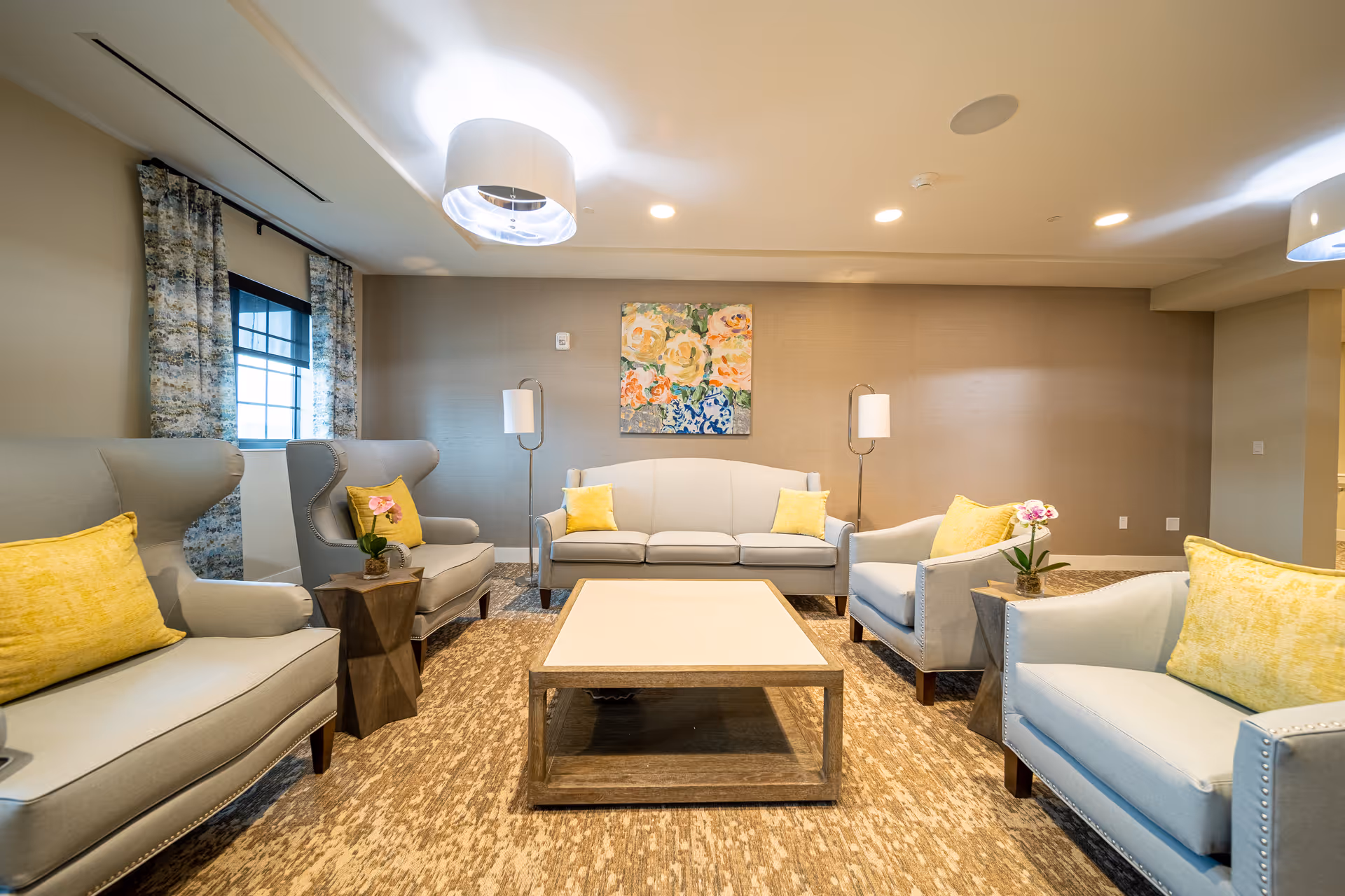 Bright, modern lounge with sofas and armchairs arranged around a central coffee table, accented by yellow pillows and wall art.