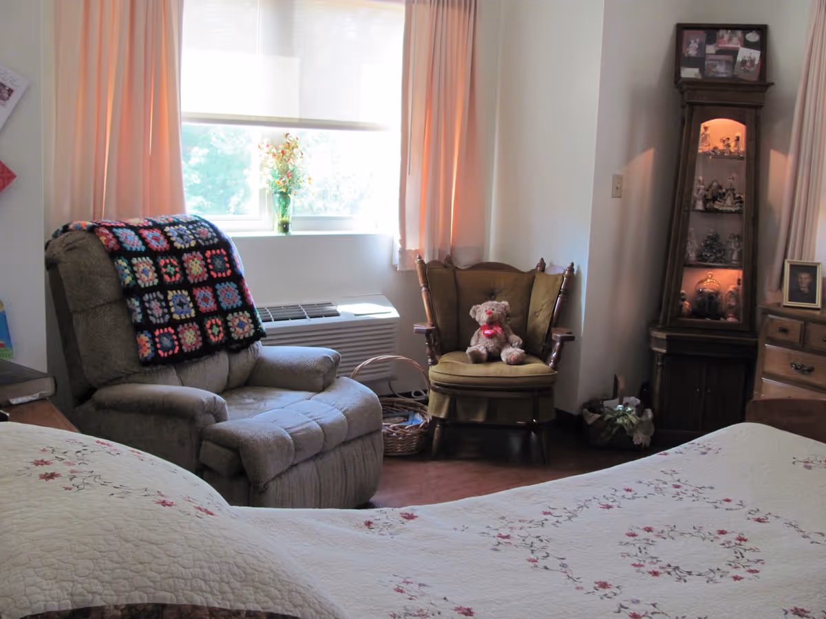 A cozy bedroom corner with a beige recliner chair draped with a colorful crocheted blanket, a wooden armchair with a teddy bear sitting on it, a window with peach-colored curtains, and a wooden display cabinet with figurines and photos. The bed with a floral quilt is partially visible in the foreground.