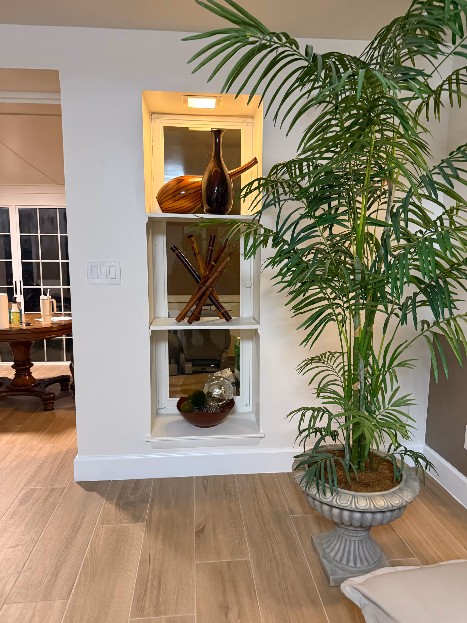 Interior view of a room with light wood flooring, a large potted plant in a decorative stone planter on the right, and a built-in wall shelf with three sections displaying decorative items including vases and wooden sticks. In the background, a wooden dining table with chairs is partially visible through an open doorway.