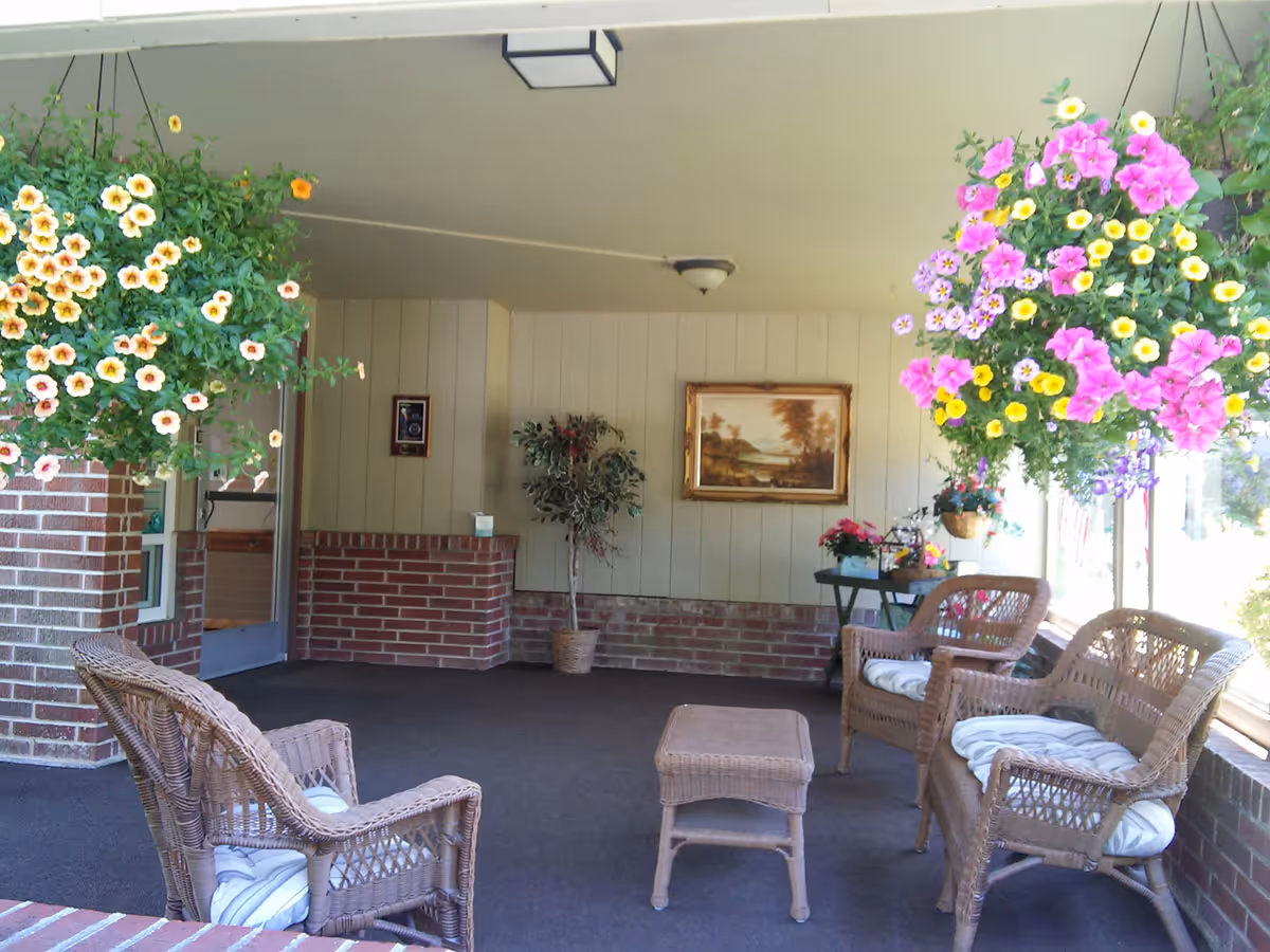 A cozy sitting area with wicker furniture and hanging flower baskets in a senior living facility.