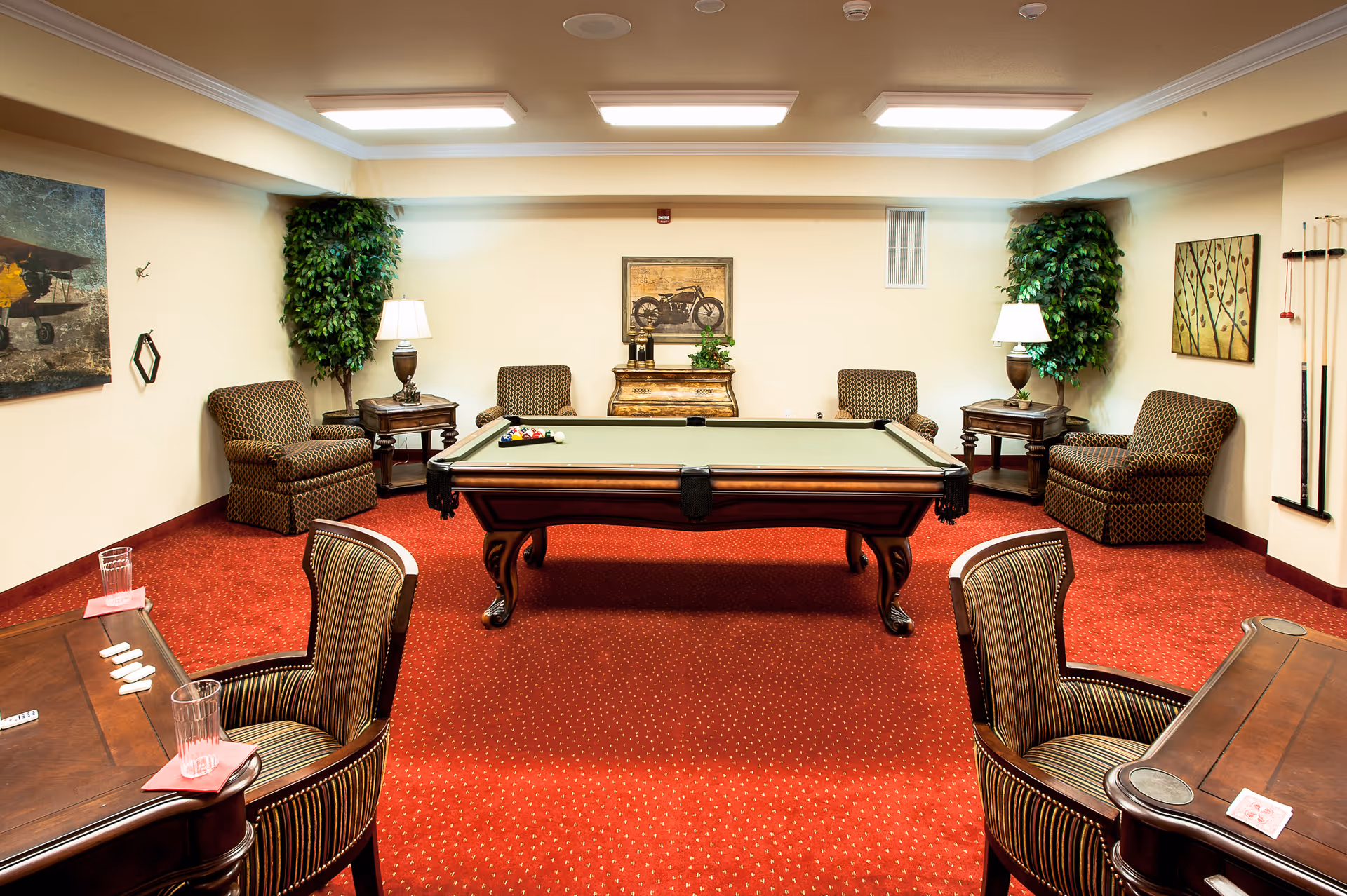A cozy game room with a pool table in the center, surrounded by four patterned armchairs and two small tables with lamps. The room has red carpet flooring, beige walls adorned with paintings, and two artificial plants in the corners. There are also two wooden tables with chairs in the foreground, each with a glass of water and playing cards.