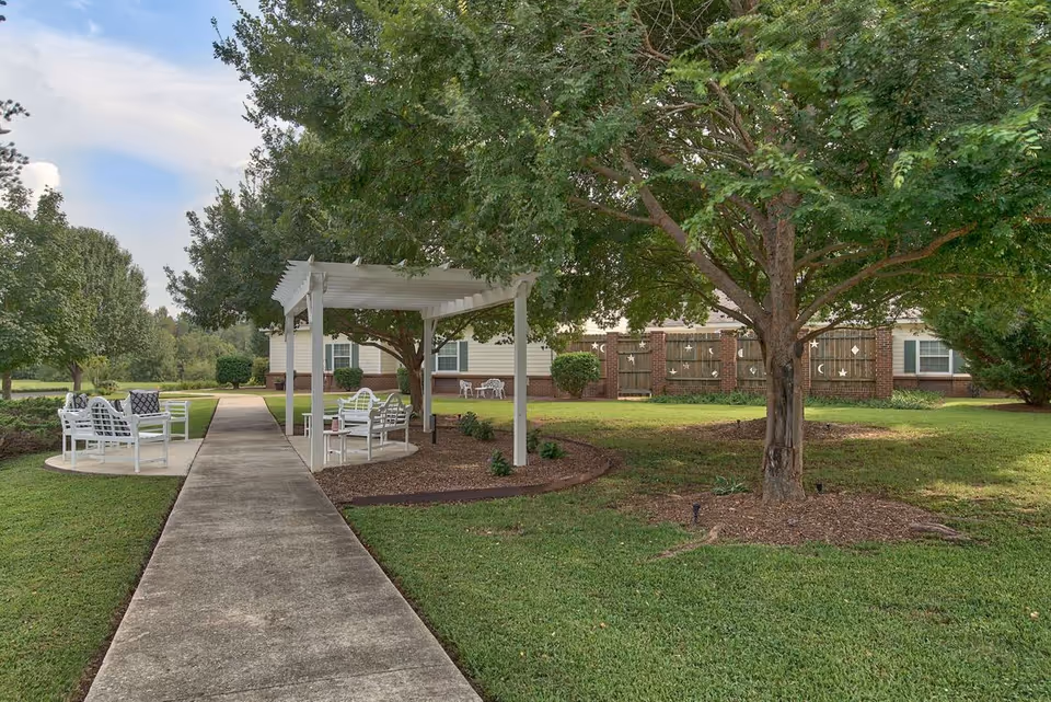 A peaceful outdoor area at Autumn Cove Assisted Living featuring a concrete walkway leading to a white pergola with white benches and chairs underneath. The area is surrounded by green grass, trees, and shrubs, with a building and decorative wooden fence in the background.