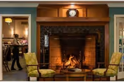 A cozy living room area featuring a lit fireplace with a wooden mantel and a clock above it. Two green patterned armchairs face each other with a small round wooden table between them. Through an open doorway, a dining area is visible with a person walking and another person seated at a table under a hanging lamp.