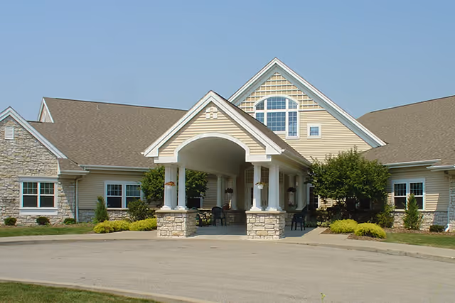 Front entrance of a beige building with a covered porte-cochère supported by columns and surrounding landscaping.