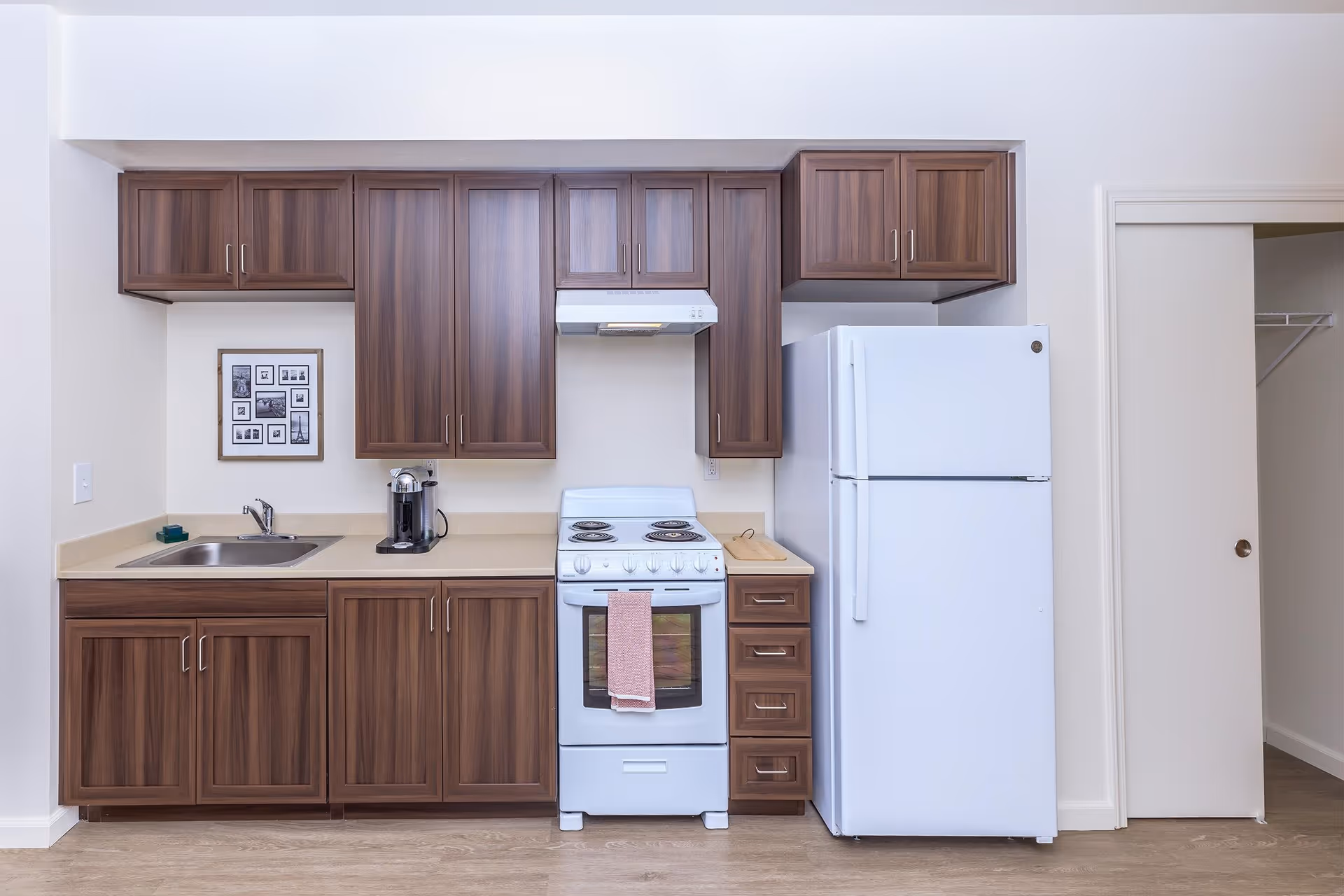 A clean and modern kitchen featuring dark wood cabinets, a white electric stove with a pink towel hanging on the handle, a white refrigerator, a stainless steel sink, a coffee maker on the countertop, and a framed collage of black and white photos on the wall.