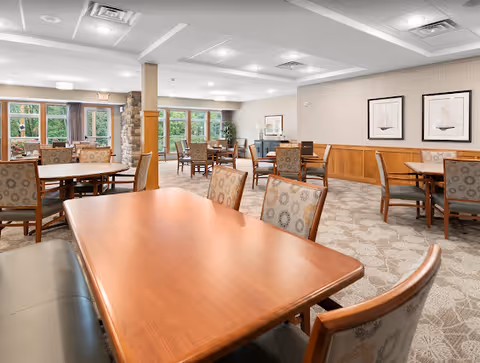 Spacious senior living dining room with multiple wooden tables and upholstered chairs, large windows, and neutral decor.