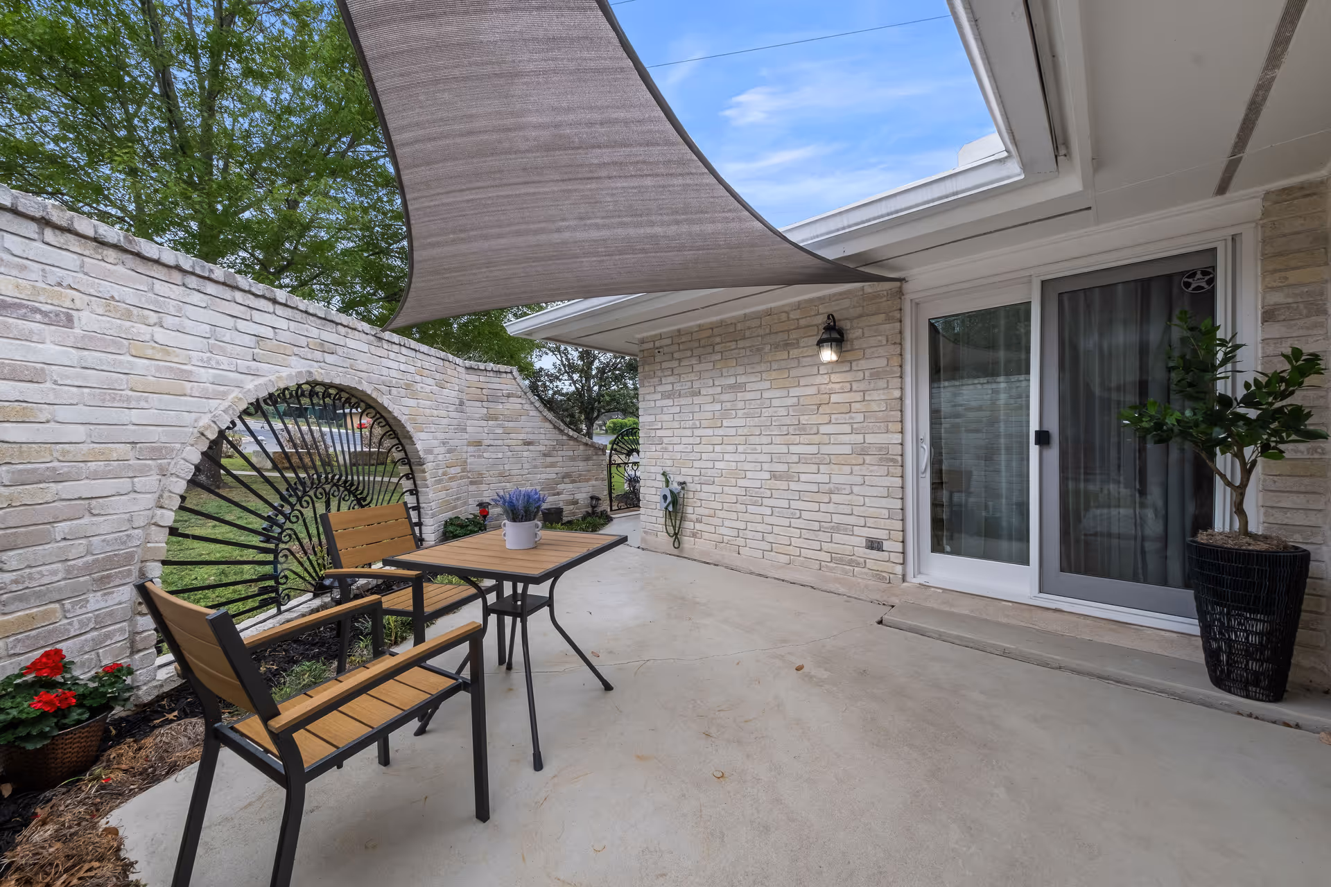 A small outdoor patio area with a table and two chairs, a potted plant on the table, and a large shade sail overhead. The patio is enclosed by a light-colored brick wall with decorative wrought iron inserts. There is a sliding glass door leading inside the building and a potted plant near the door.