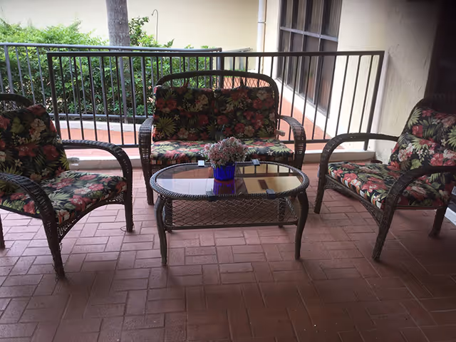 Outdoor patio area with a set of wicker furniture including two armchairs and a loveseat, all with floral patterned cushions, and a glass-top coffee table with a small blue vase holding flowers. The patio has a brick floor and is enclosed by a metal railing with greenery visible beyond.