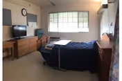 A simple, clean bedroom in a senior living facility with a single bed covered in a dark blue blanket, a wooden dresser with a flat-screen TV on top, a chair, a wall clock, and a large window letting in natural light.