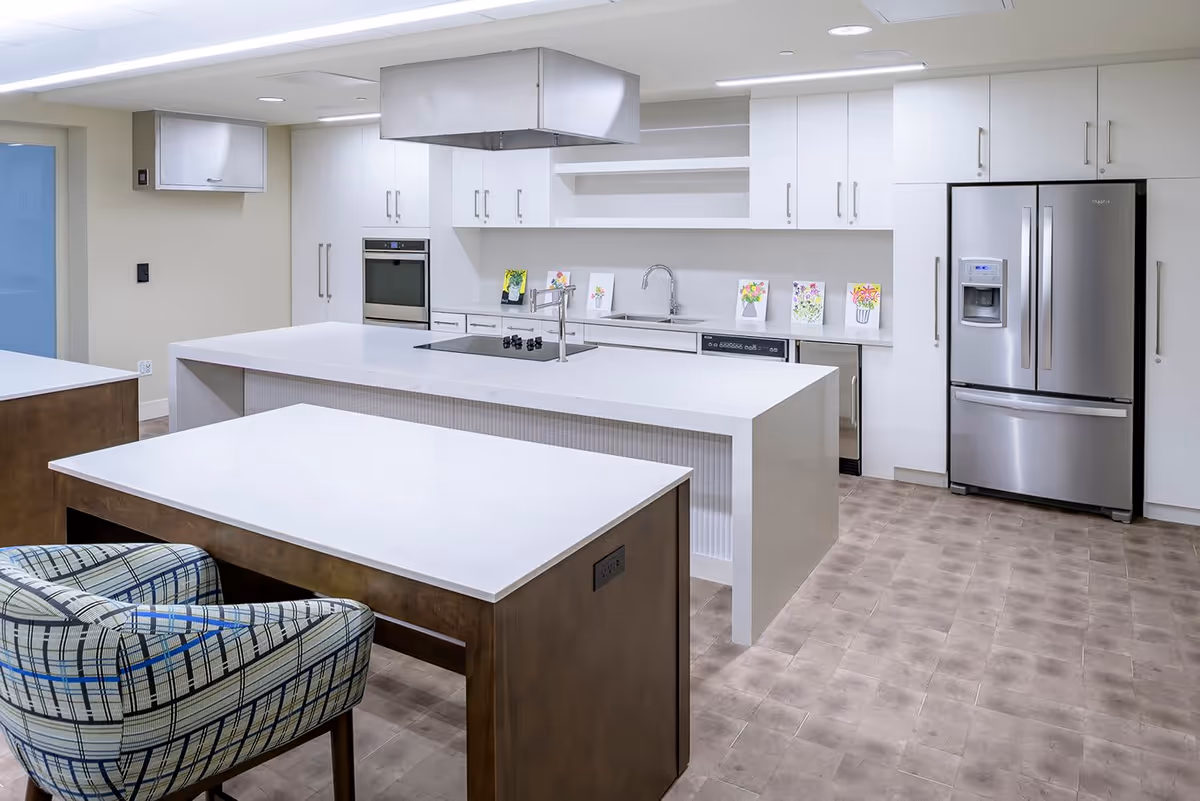 Modern senior living facility kitchen with white countertops, stainless steel refrigerator, built-in oven, stovetop with overhead vent, and a patterned armchair at a wooden table.