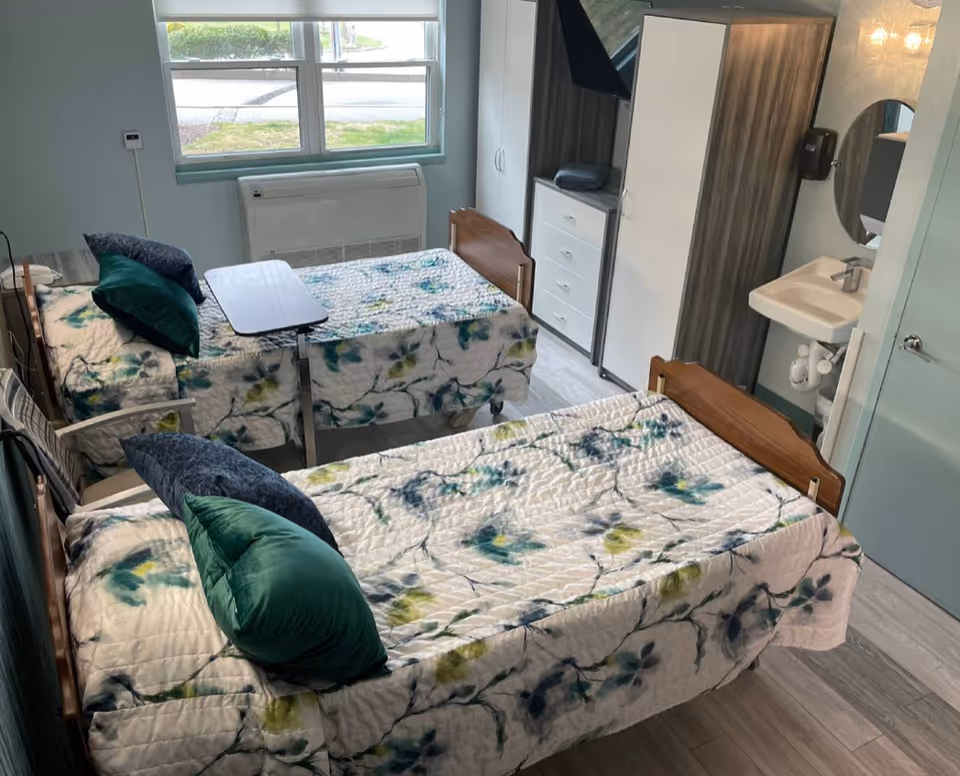 A bright shared bedroom with two twin beds covered in floral quilts, green pillows, an overbed table, wardrobe, sink, and a window with an air unit.