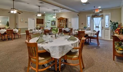 A spacious dining room with several wooden tables and chairs arranged neatly. Each table is covered with a white tablecloth and set with plates, cups, and green napkins. The room has beige walls, carpeted floors, ceiling fans with lights, and large windows with curtains allowing natural light to enter. There are decorative plants and a wooden cabinet along the walls.