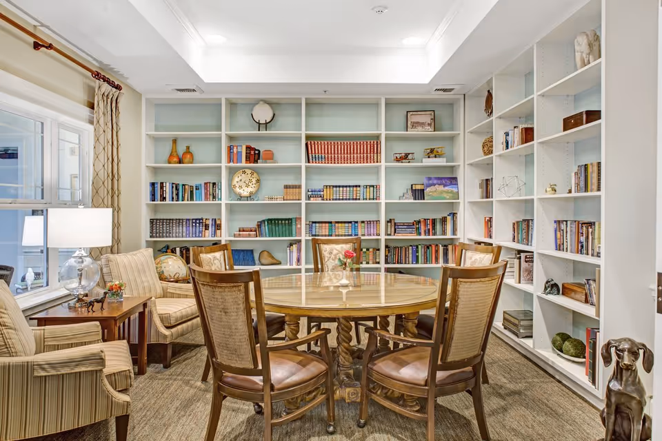 A cozy reading or sitting room with a round wooden table surrounded by four wooden chairs with cushioned seats. Behind the table, there is a large white built-in bookshelf filled with books and decorative items. To the left, there are two striped armchairs with a wooden side table between them, holding a lamp and a small flower arrangement. The room has a window with beige curtains and a skylight above, providing natural light.