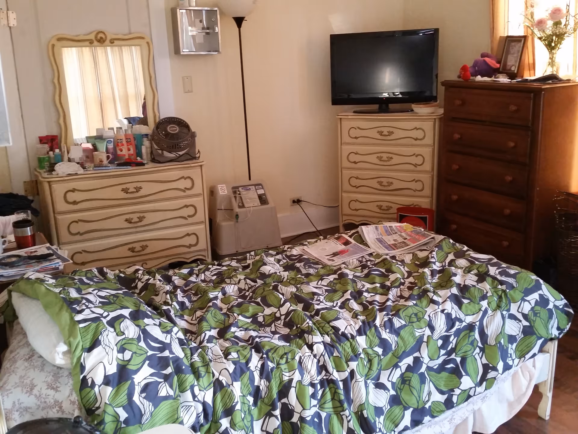 A bedroom with a bed covered in a floral patterned comforter in green, white, and navy blue. There are two dressers, one cream-colored with ornate handles and a mirror, and another wooden dresser with a TV on top. Various personal items and papers are scattered on the bed and dressers. A floor lamp and a small appliance are also visible in the corner.