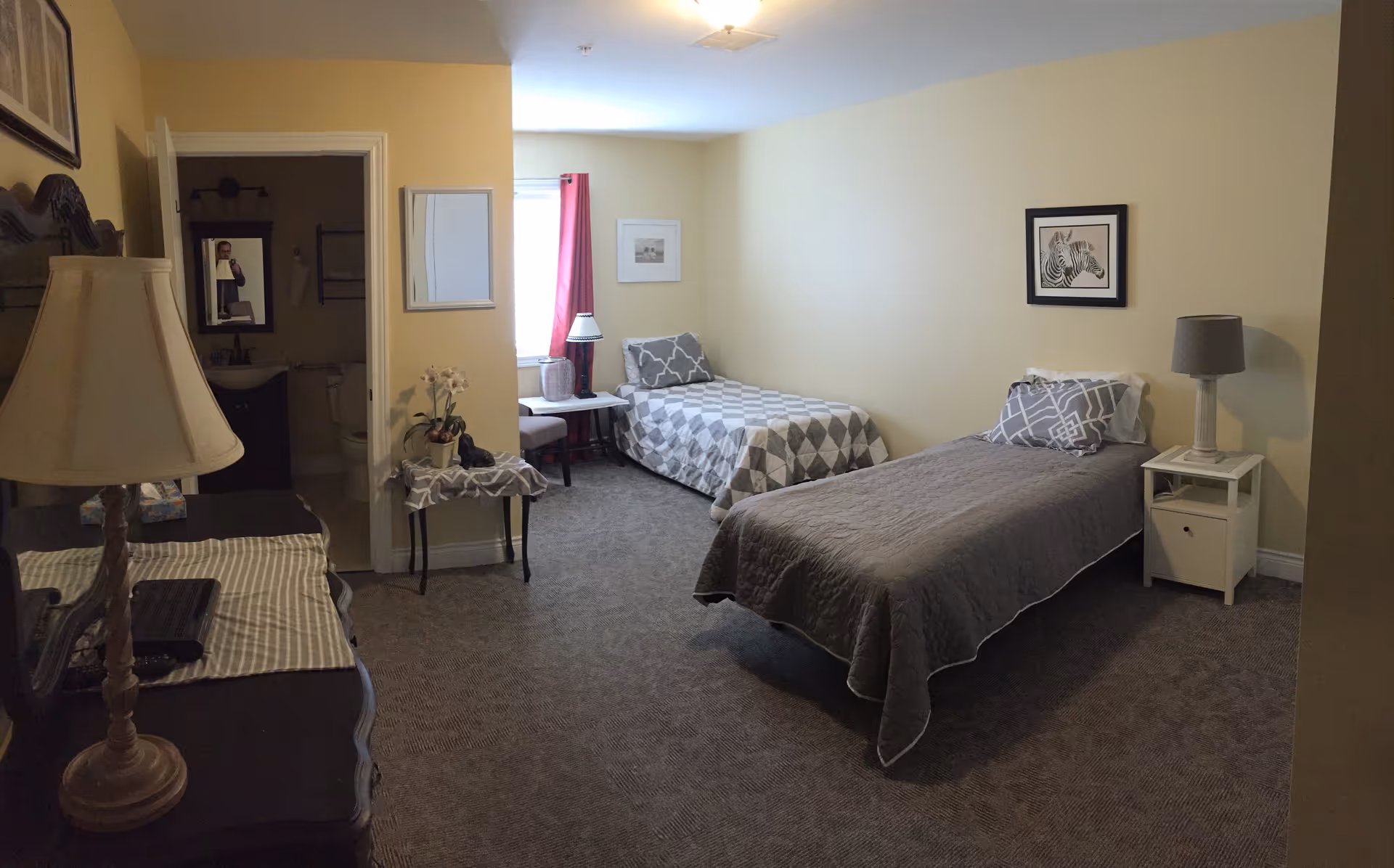 A bedroom with two single beds, each with gray and white bedding and pillows. There is a small white nightstand with a lamp next to one bed, and a small table with a lamp and chair near the window with red curtains. A dresser with a lamp and some items on top is on the left side of the room. The walls are painted light yellow, and there is a framed picture of a zebra on the wall. An open door reveals a bathroom with a sink and toilet.