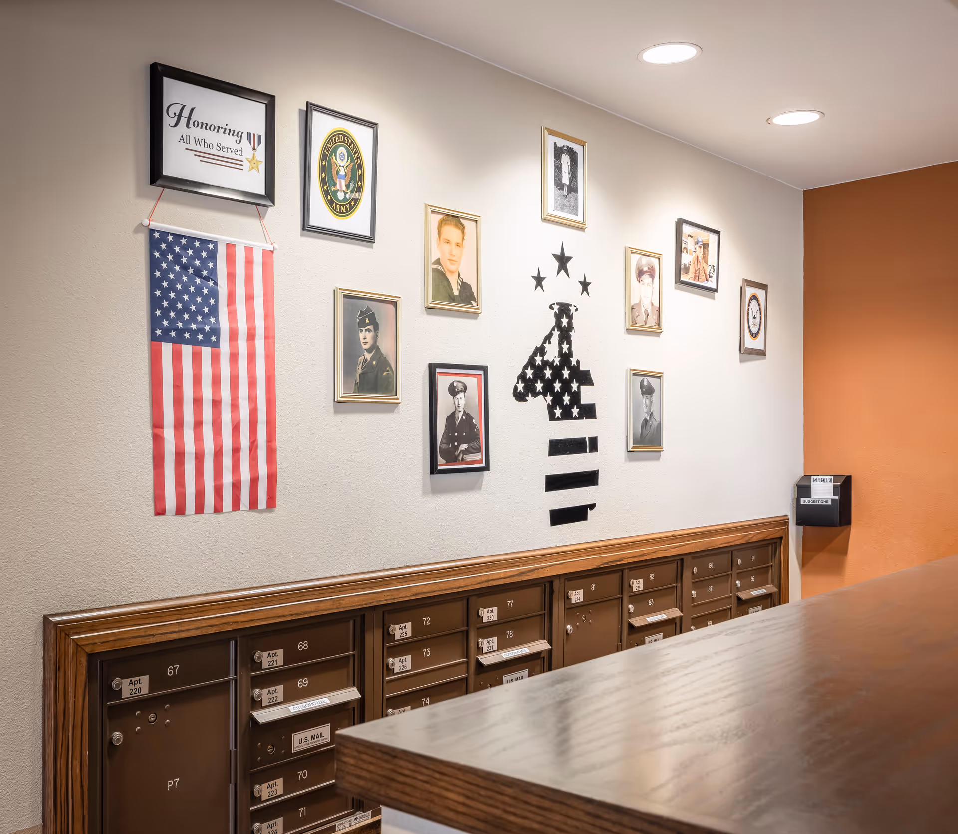 Interior view of a mailroom area with multiple mailboxes labeled with apartment numbers. Above the mailboxes, there is a wall decorated with framed photos of military personnel, a United States Army emblem, an American flag hanging vertically, and a sign that reads 'Honoring All Who Served'. The wall also features a black silhouette of a soldier with stars above it. The room has a wooden countertop in the foreground and an orange accent wall on the right side.