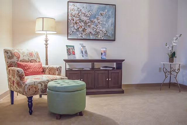 A cozy living area with a patterned armchair featuring a red cushion, a green round ottoman, a wooden cabinet with brochures on top, a floor lamp, a floral painting on the wall, and a small side table with a potted plant.