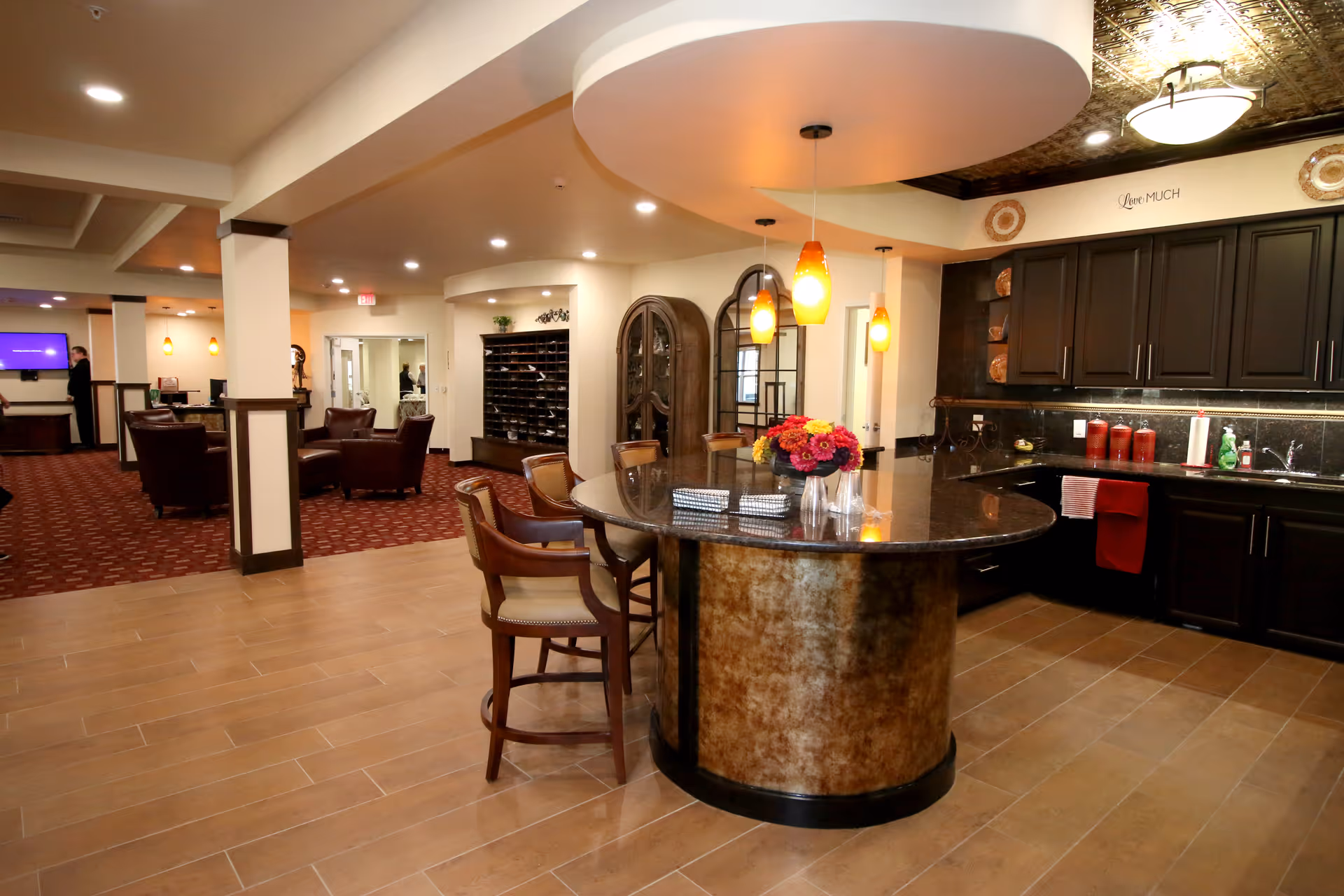 A spacious interior area featuring a kitchen with dark wood cabinets, a large curved island with a granite countertop, and three pendant lights hanging above. The island has chairs around it and a vase with colorful flowers on top. In the background, there is a seating area with leather armchairs and a carpeted floor, as well as a wall with cubby shelves and a decorative cabinet.