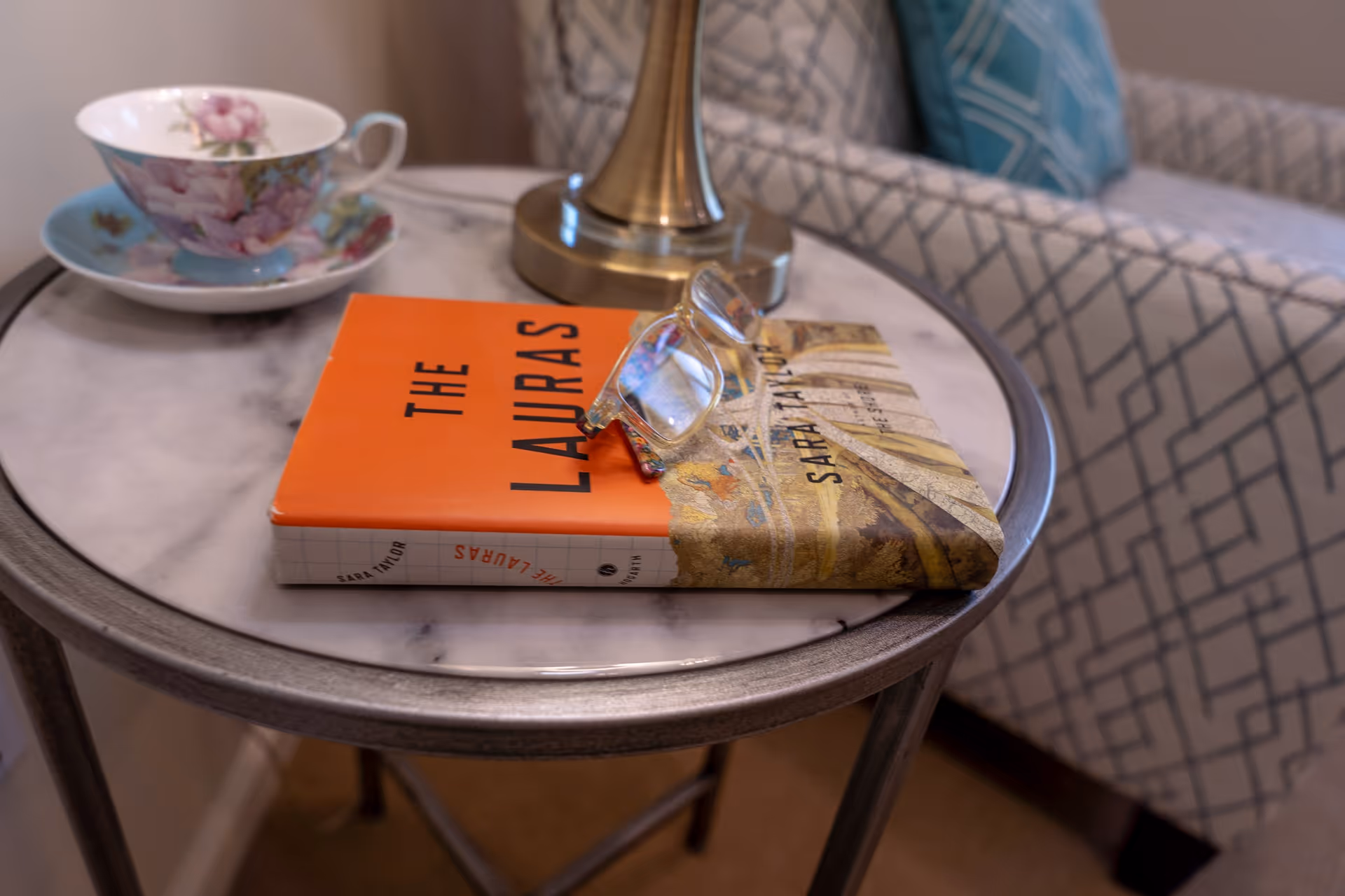 A small round side table with a marble top holding a floral teacup and saucer, a brass lamp, a book titled 'The Lauras' by Sara Taylor, and a pair of reading glasses. In the background, there is a patterned armchair with a blue cushion.