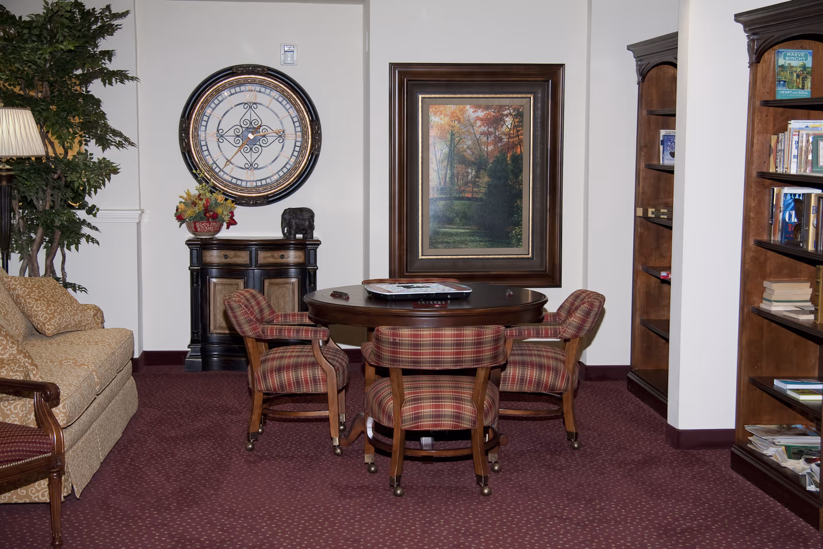 Cozy senior living common area with a round wooden table surrounded by four plaid upholstered chairs on casters. A large decorative wall clock and a framed autumn landscape painting hang on the wall. To the left, there is a beige patterned sofa and a tall green plant with a floor lamp. On the right, wooden bookshelves filled with books are visible.