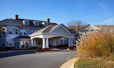 Front entrance of a multi-story senior living building with a covered porte-cochere supported by white columns and ornamental grasses by the driveway.