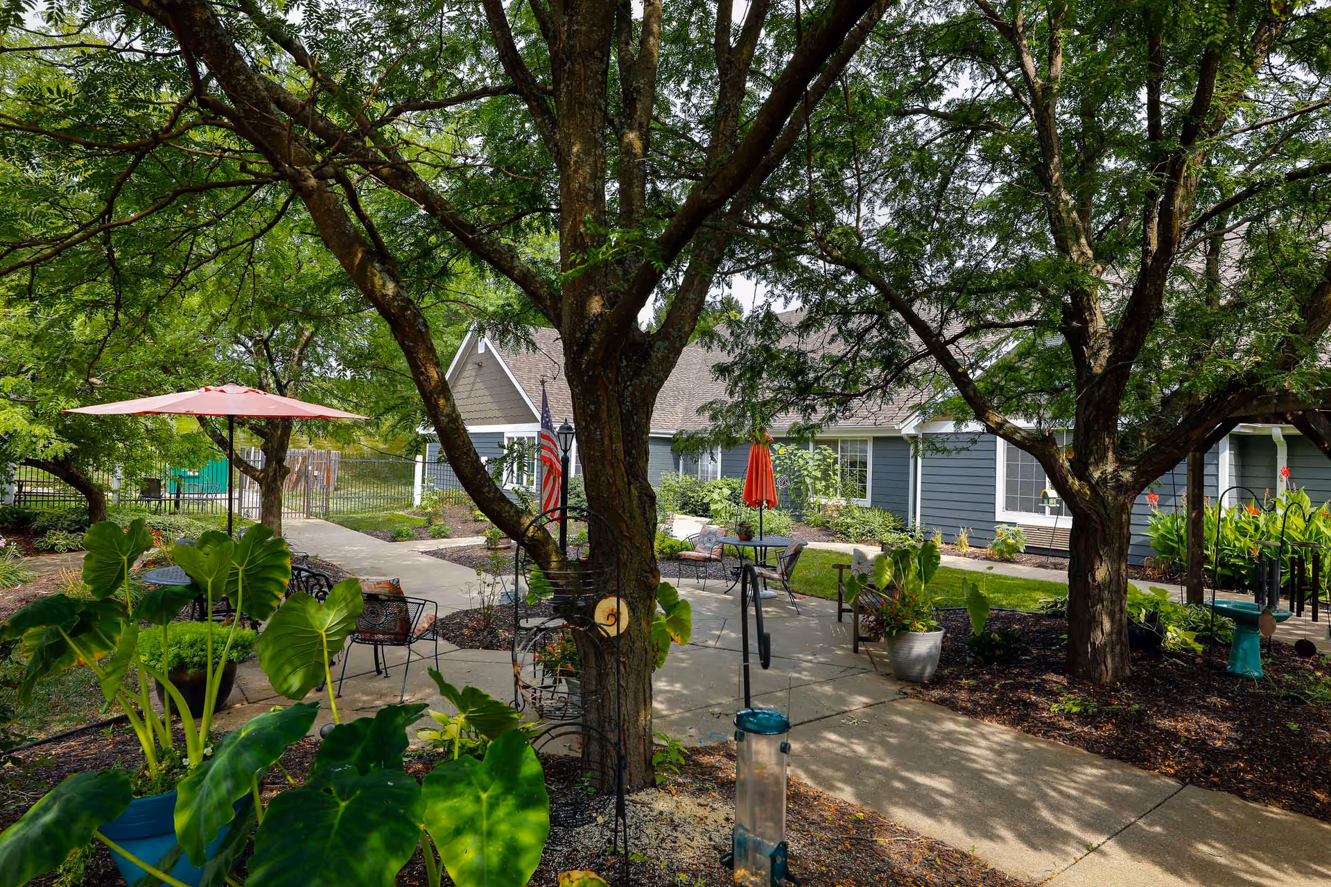 Outdoor garden area at Homestead Assisted Living of Leavenworth featuring large trees providing shade, potted plants, a paved walkway, several seating areas with chairs and tables, and umbrellas. The building with blue siding and white trim is visible in the background.