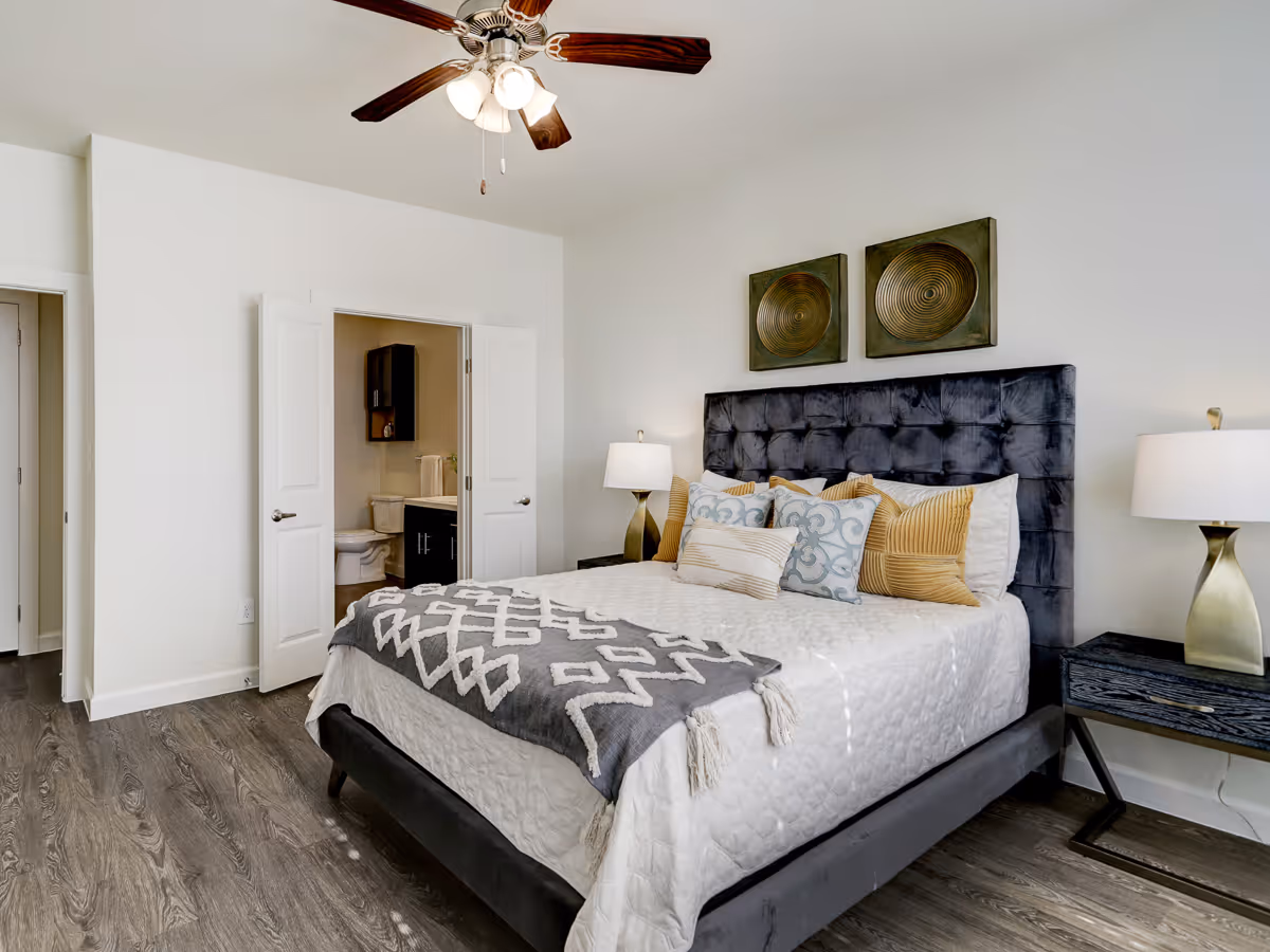 A modern bedroom with a large bed featuring a tufted dark gray headboard, white bedding, and multiple decorative pillows. There are two bedside tables with matching lamps on either side of the bed. The room has wood flooring, white walls, and a ceiling fan with lights. An open door reveals a bathroom with a toilet and vanity.