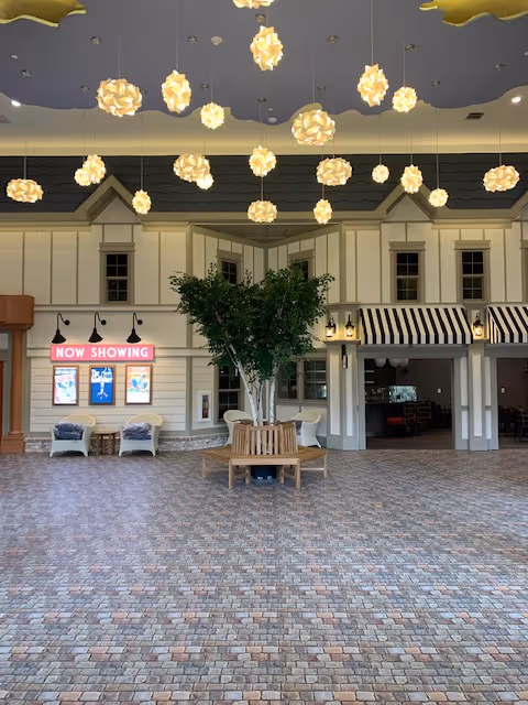 Spacious indoor common area with a cobblestone-patterned floor, a central tree surrounded by a circular wooden bench, hanging flower-shaped pendant lights from the ceiling, and a facade resembling storefronts with windows and awnings. A 'NOW SHOWING' sign with movie posters is visible on the left side.