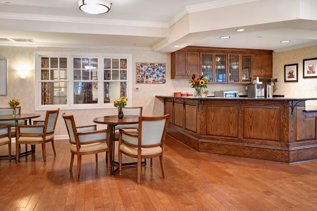 A cozy common area with wooden floors, round tables with chairs, and a wooden counter with cabinets. The counter has a coffee machine, a microwave, and a vase with sunflowers. The walls are decorated with framed pictures and a collage of photos. There is a window with white trim and patterned curtains visible through it.