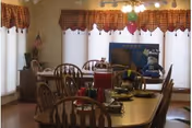 A dining room with wooden tables and chairs arranged around them. The tables have pitchers, cups, and plates with food. The room has large windows covered with white curtains and red valances. There are balloons hanging from the ceiling and an American flag in the corner.