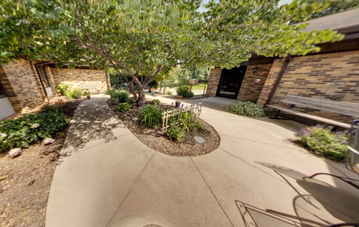 Outdoor courtyard area with a concrete walkway curving around a small garden bed with plants and a tree. There are brick buildings on either side with benches along the walls and greenery surrounding the path.