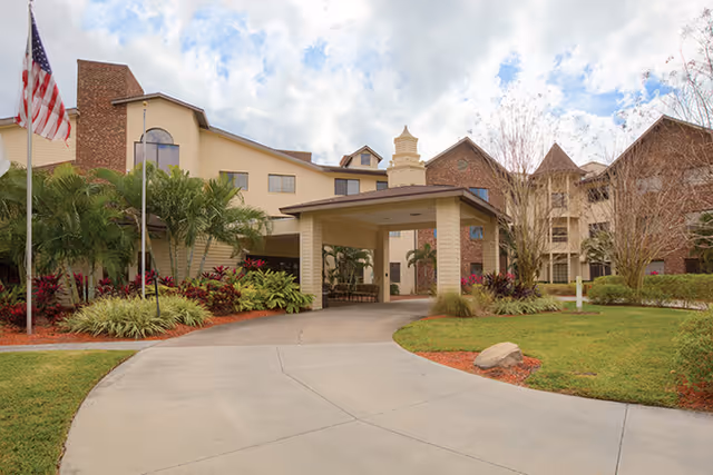 Exterior view of Freedom Village of Bradenton showing a multi-story building with a covered entrance, landscaped greenery, palm trees, and an American flag on a flagpole under a partly cloudy sky.