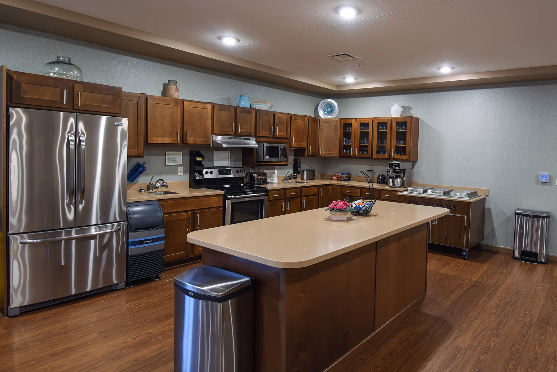 Large communal kitchen with a central island, stainless steel appliances, wooden cabinets and hardwood floors.