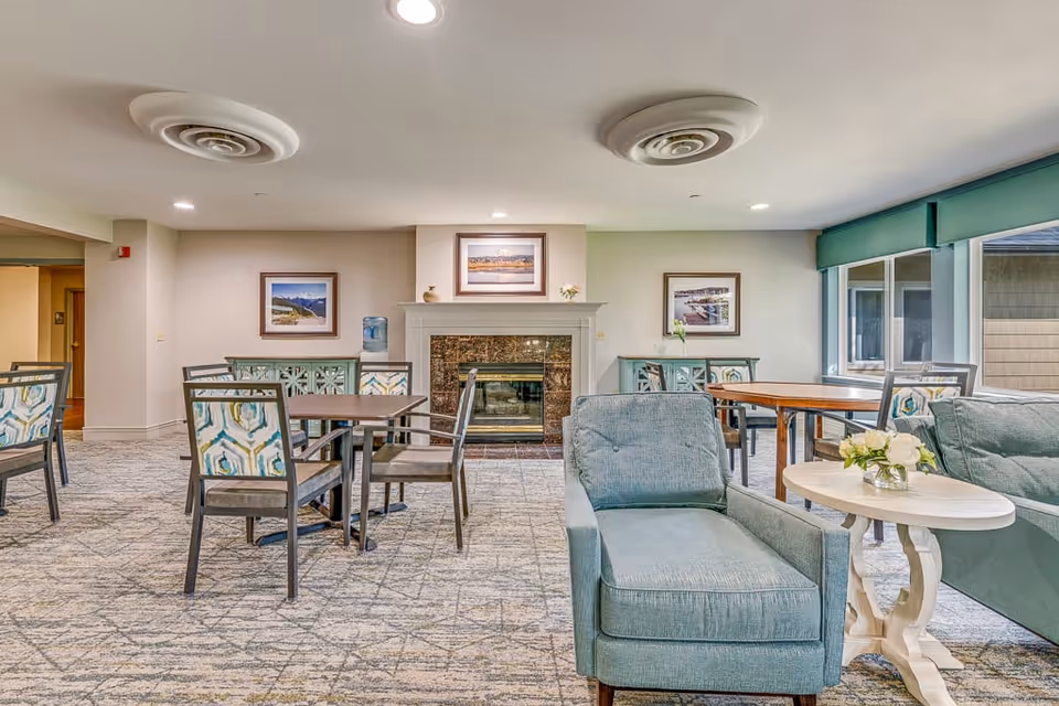 A senior living facility common area with multiple tables and chairs arranged for seating. There is a fireplace with framed pictures above it on the far wall. Two blue upholstered armchairs and a small white round table with a flower arrangement are in the foreground. Large windows on the right side let in natural light.