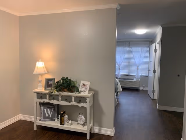 Interior view of a senior living facility showing a small white console table with decorative items including a lamp, framed photos, a plant, books, and a clock. The table is against a beige wall with dark wood flooring. In the background, there is a hallway leading to a room with a bed, window with white curtains, and an air conditioning unit below the window.