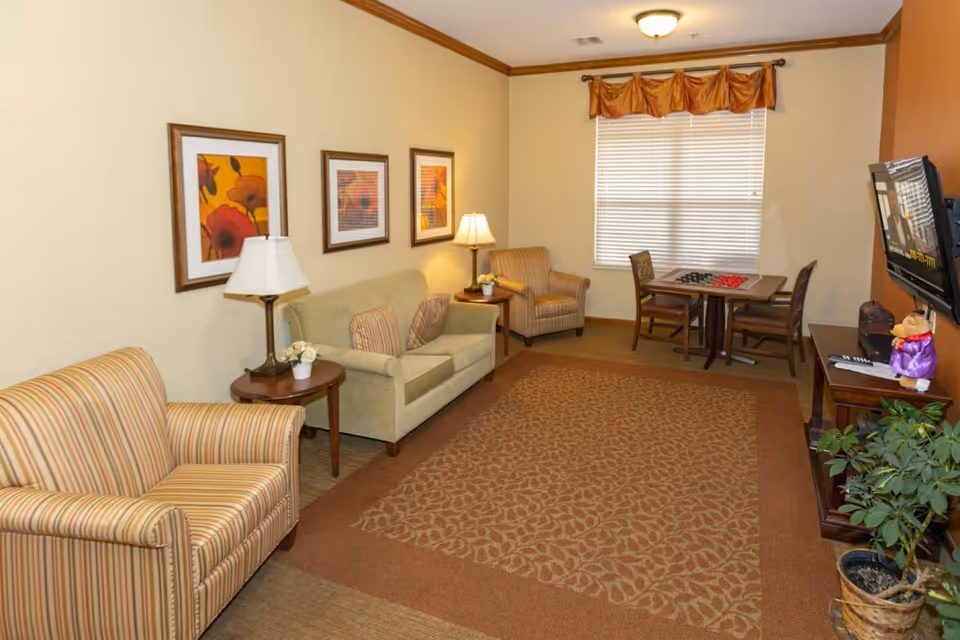 Comfortable sitting area with armchairs, a sofa, side tables, framed artwork, a TV, and a small game table by a window.