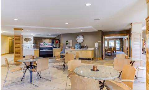Bright communal dining area with round tables and chairs, a beverage/snack station and wall clock on the back wall.