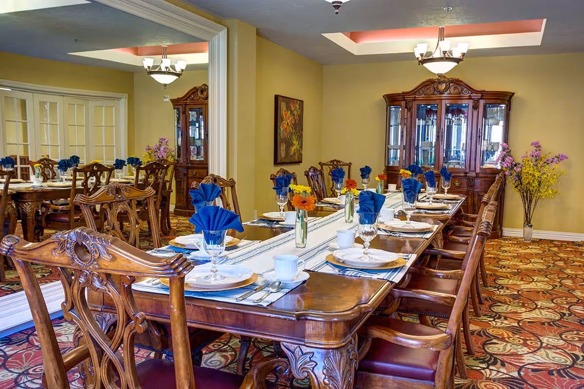 A formal dining room with a long wooden table set for a meal. The table is adorned with white plates, glassware, silverware, blue folded napkins, and small vases with colorful flowers. The room features ornate wooden chairs, a patterned carpet, a large wooden china cabinet, and a ceiling light fixture. A large mirror on one wall reflects part of the room.