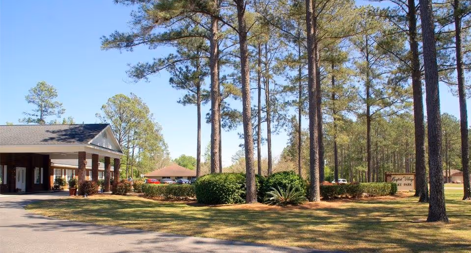 Exterior view of Baptist Village Lake Park facility showing a building with a covered entrance, surrounded by tall pine trees, bushes, and a well-maintained lawn under a clear blue sky.