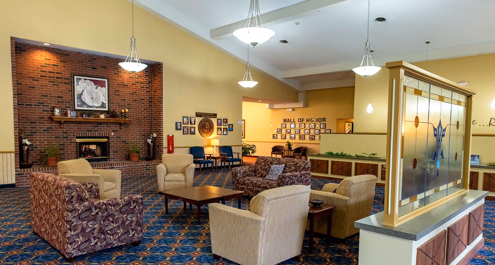 A spacious living room area in a senior living facility featuring a brick fireplace with a floral painting above it. The room has patterned carpet flooring and is furnished with multiple armchairs and sofas arranged around a central wooden coffee table. There are hanging pendant lights from the ceiling, and a decorative stained glass partition on the right side. In the background, a wall labeled 'Wall of Honor' displays framed photos.