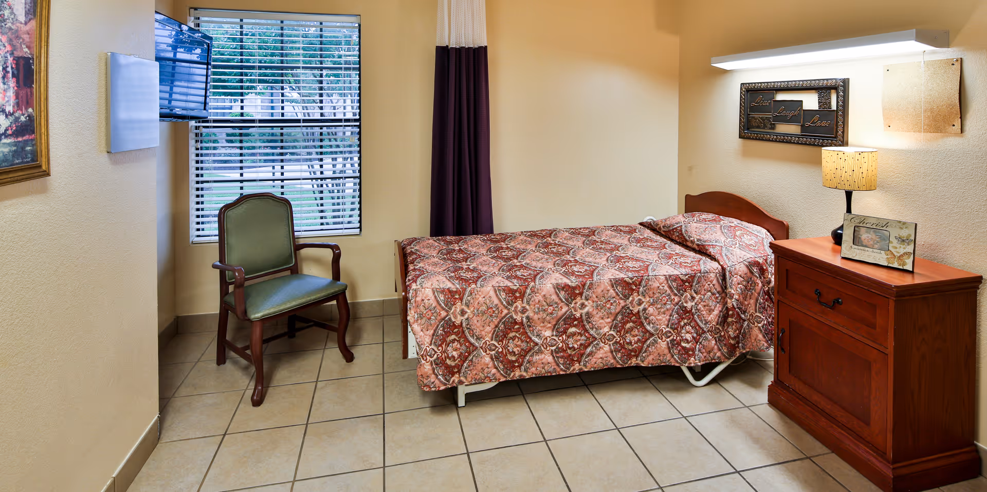 Small furnished bedroom with a single bed covered by a patterned quilt, a wooden nightstand with a lamp, and a chair by a window.