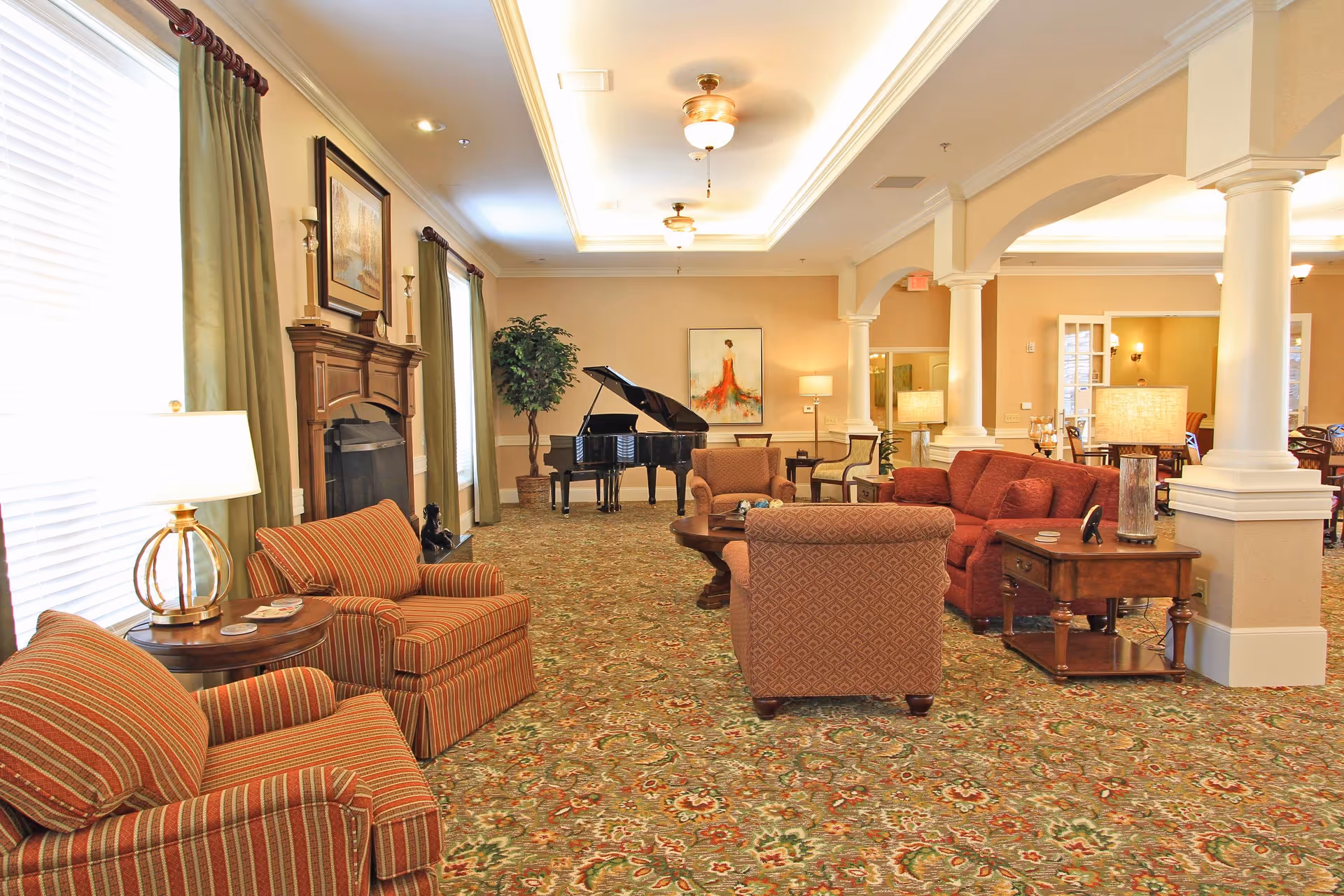 A spacious and well-lit living room area in Benton House of Staley Hills featuring patterned carpet, multiple upholstered chairs and sofas in warm tones, a wooden coffee table, side tables with lamps, a grand piano near the back wall, a fireplace with framed artwork above it, tall windows with green curtains, and decorative columns separating the seating area from an adjoining dining space.