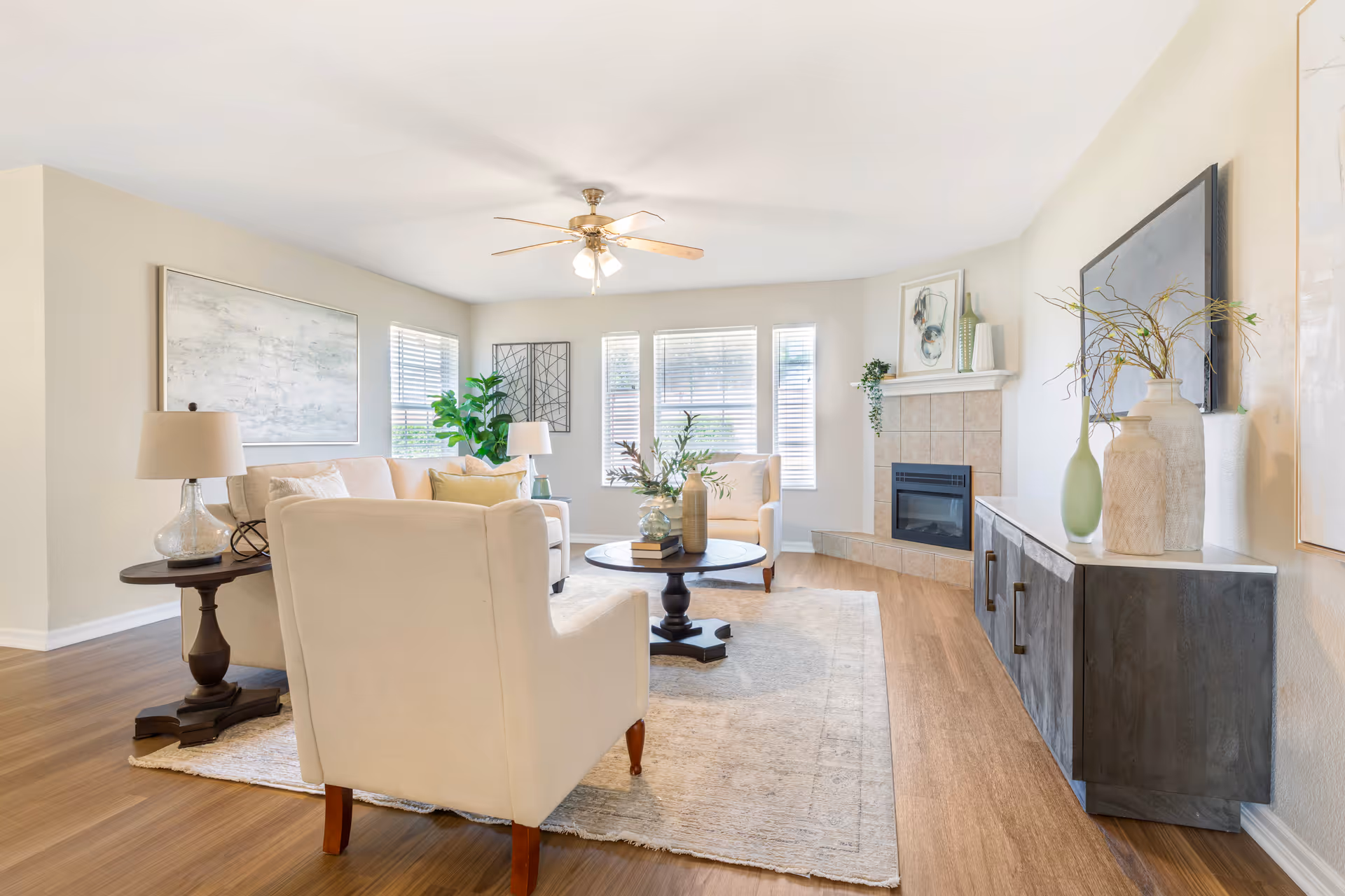 Bright furnished living room with cream chairs and sofa around a round coffee table, a fireplace, and large windows.