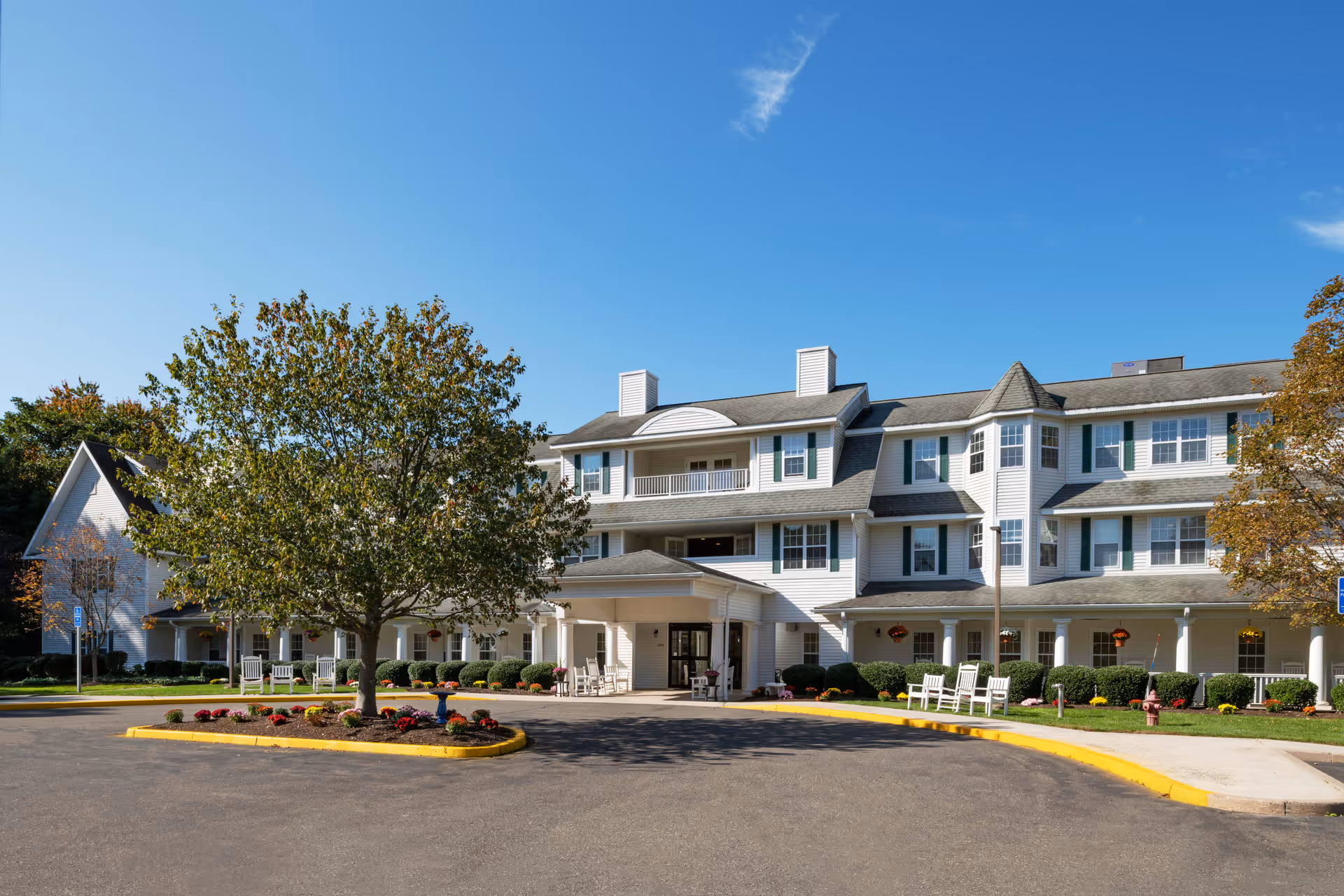 Exterior view of a large, multi-story senior living facility building with white siding, green shutters, and a covered entrance. There is a circular driveway with a landscaped island featuring a tree and flowers. Several white rocking chairs are placed along the front porch. The sky is clear and blue.