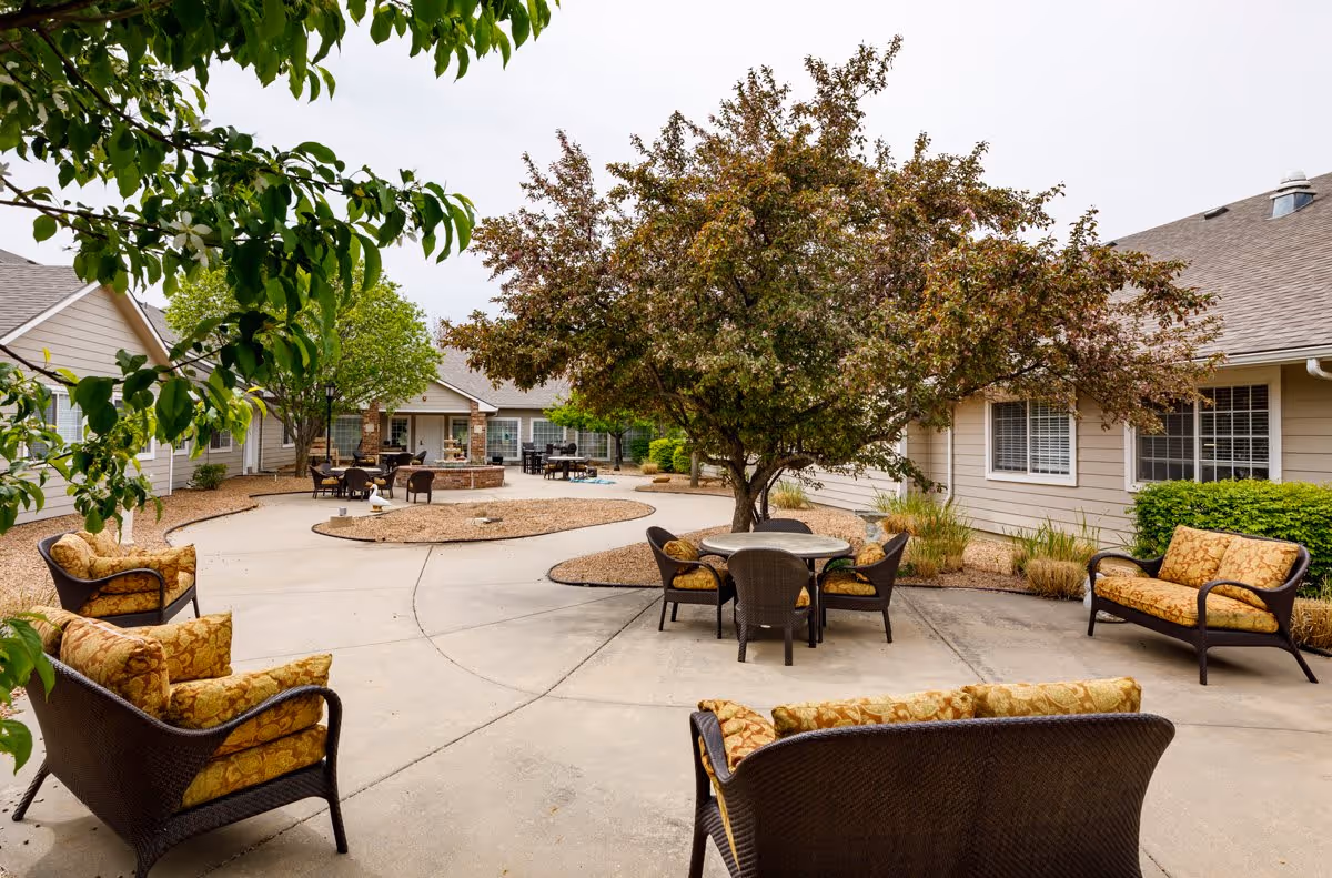 Outdoor courtyard area at Homestead Estates of Wichita featuring cushioned wicker seating arranged around a concrete patio with a tree in the center and additional seating near a fire pit in the background, surrounded by single-story beige buildings with windows and greenery.