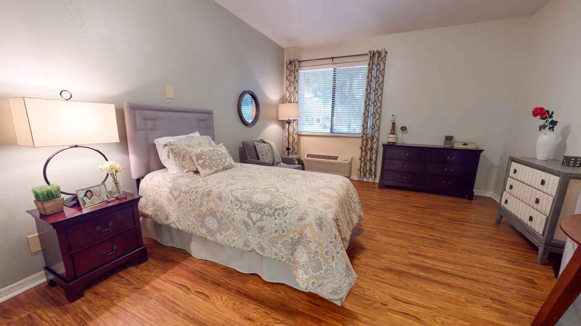 A neatly arranged bedroom with a single bed covered in a patterned beige and white bedspread. There is a wooden nightstand with a lamp, a small plant, and a framed photo next to the bed. A round mirror hangs on the wall above a gray armchair near a window with patterned curtains. Across the room, there are two dressers, one dark wood and one lighter wood with a vase holding a red rose. The floor is wooden and the walls are light-colored.