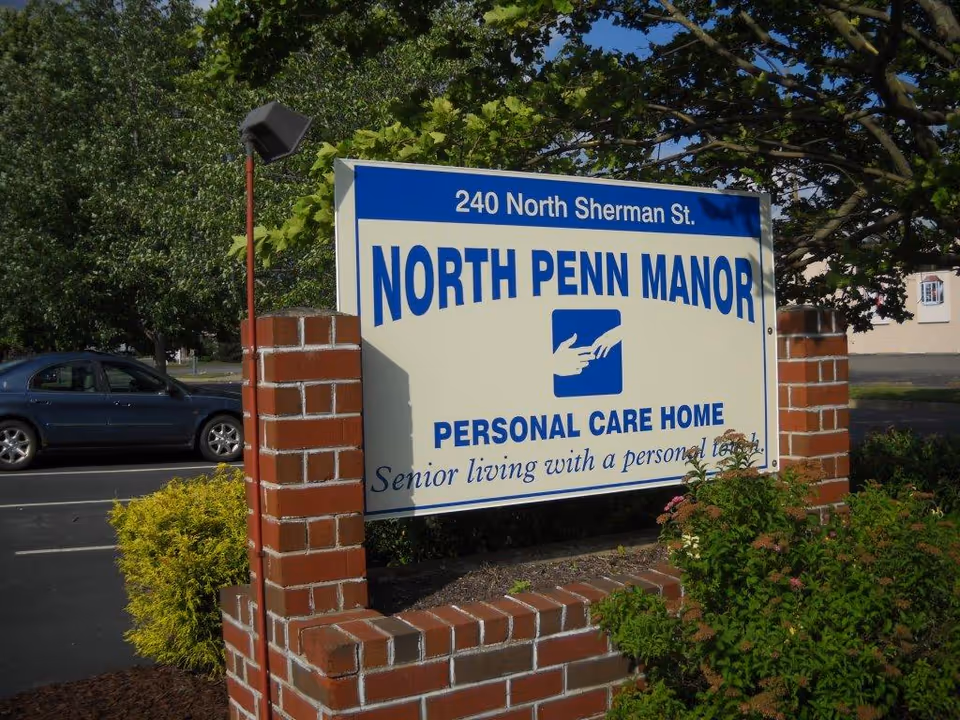 Outdoor sign for North Penn Manor Personal Care Home located at 240 North Sherman St., surrounded by greenery and a brick frame, with a parked car and trees in the background.