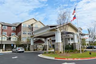 Exterior view of Ashwood Meadows Gracious Retirement Living facility showing a multi-story building with a covered entrance driveway, landscaped grounds, and two flagpoles flying the American and Texas flags under a partly cloudy sky.